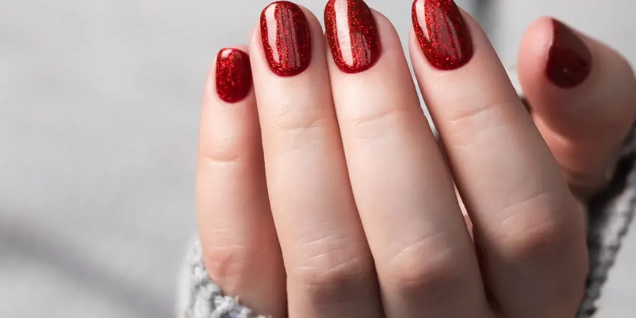 Glitzernde rote Gel-Nagel-Designs auf einer Hand, die aus einem grauen Pullover ragt.