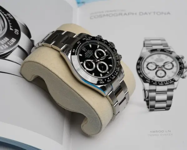 Luxuriöse Rolex Daytona Uhr auf einem Kissen vor einem Buch mit Uhrenabbildungen.
