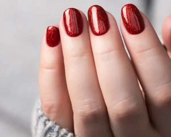 Glitzernde rote Gel-Nagel-Designs auf einer Hand, die aus einem grauen Pullover ragt.