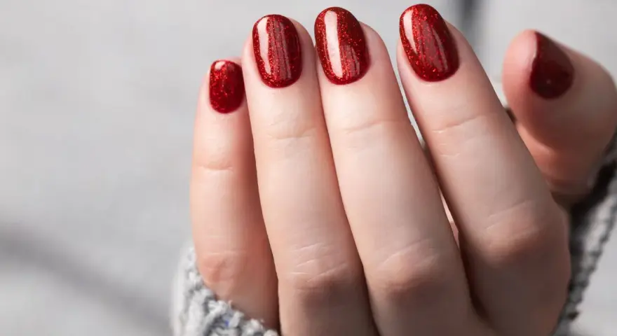Glitzernde rote Gel-Nagel-Designs auf einer Hand, die aus einem grauen Pullover ragt.
